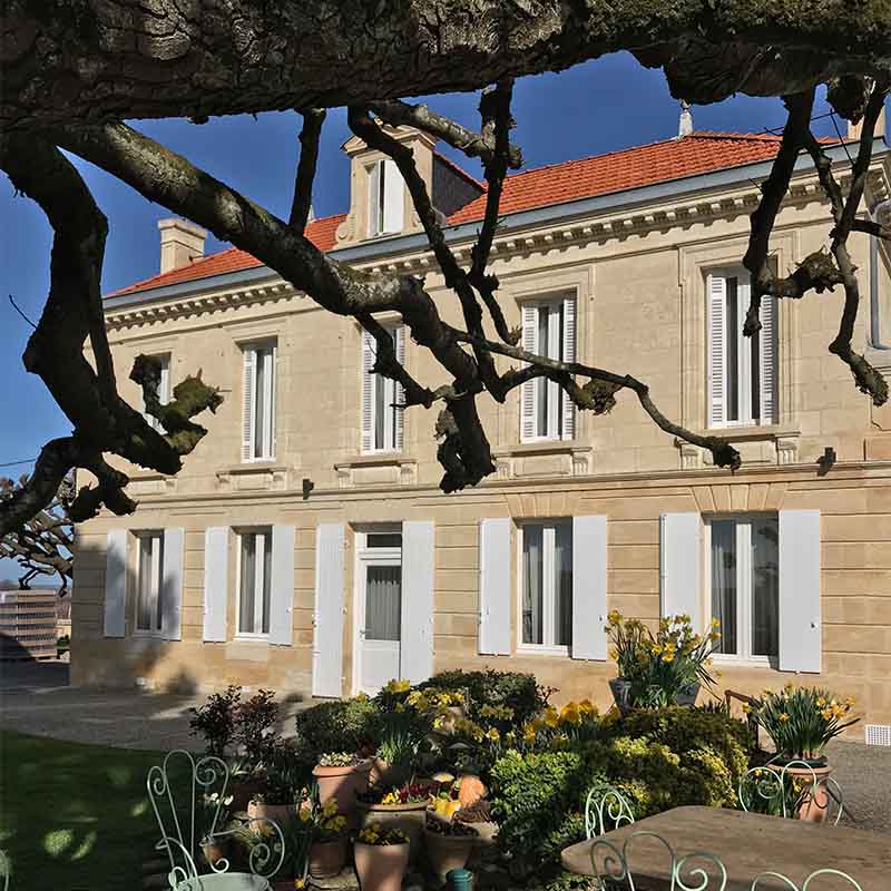 The Château Belair Coubet, a traditional stone estate with white shutters and a landscaped garden framed by mature vine branches.